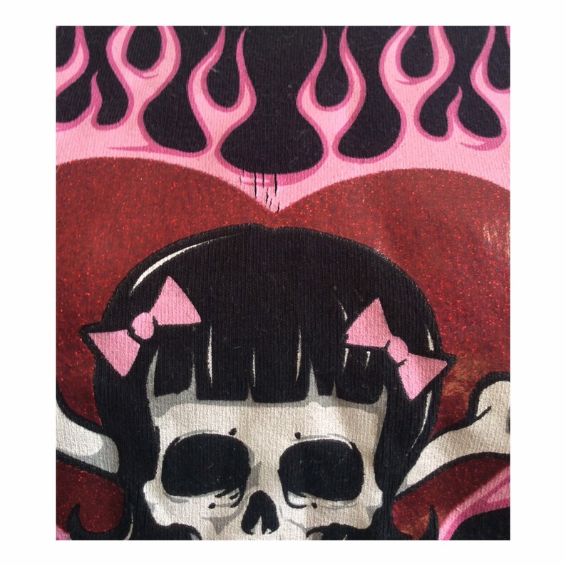 May include: A black and white skull with pink bows on the side of its head. The skull is surrounded by a red heart with pink flames behind it.