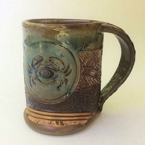 May include: A brown ceramic mug with a blue and green crab design. The mug has a textured surface and a handle.