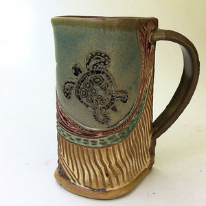 May include: A ceramic mug with a green, brown, and beige glaze. The mug features a raised design of a sea turtle with a detailed shell. The mug has a handle and a textured, striped pattern on the bottom.