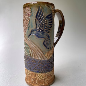 May include: A ceramic mug with a brown, blue, and green glaze. The mug features a raised design of a hummingbird in flight, surrounded by floral and geometric patterns.