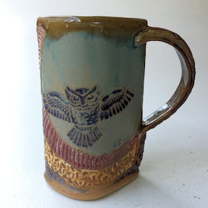 May include: A ceramic mug with a brown, blue, and red design. The mug features a stylized owl in flight with a blue and white pattern. The mug has a handle and is made by hand.