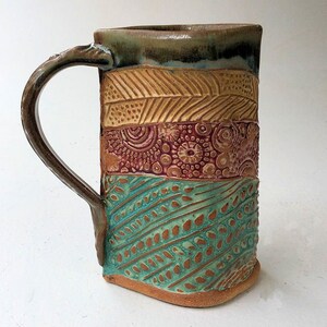 May include: A handmade ceramic mug with a brown handle and a colorful design. The mug is decorated with a pattern of brown, red, and turquoise. The mug is a unique and beautiful piece of pottery.