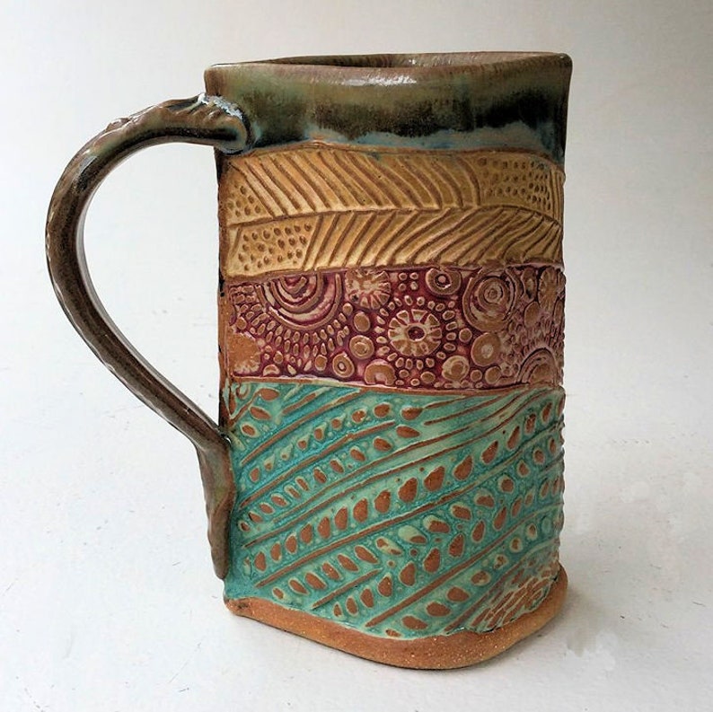 May include: A handmade ceramic mug with a brown handle and a colorful design. The mug is decorated with a pattern of brown, red, and turquoise. The mug is a unique and beautiful piece of pottery.