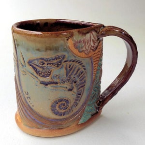 May include: A ceramic mug with a brown handle and a light blue glaze. The mug features a detailed, embossed design of a chameleon with a purple and blue color scheme.