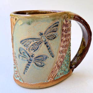 May include: A ceramic mug with a light blue glaze and a brown handle. The mug has a textured surface with a design of two dragonflies in blue and brown.