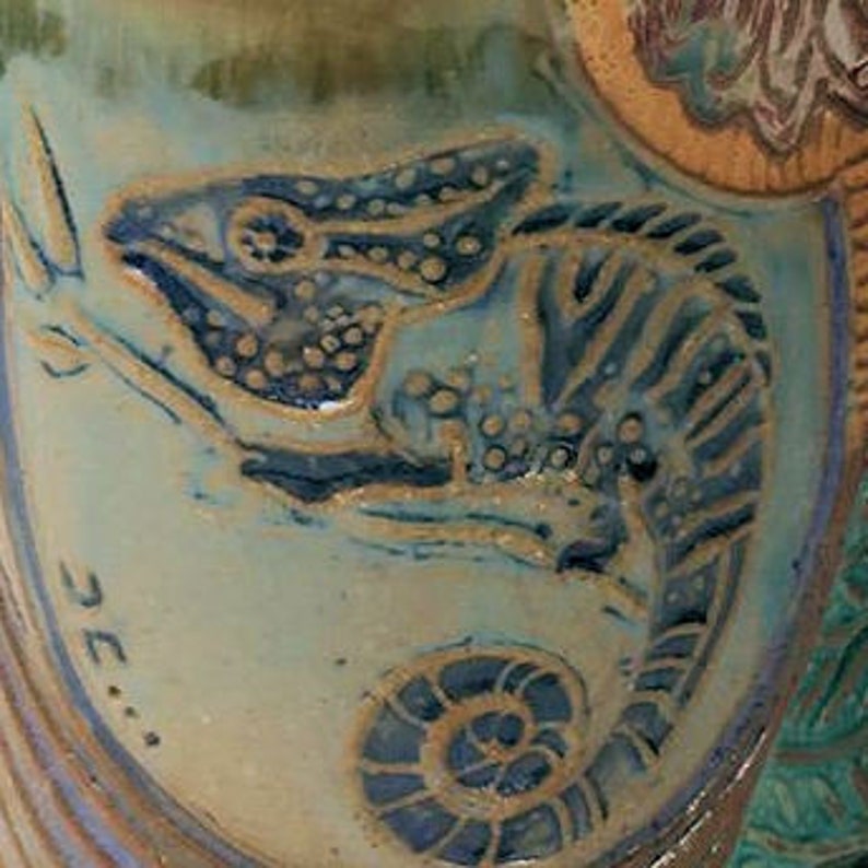 May include: A ceramic tile with a green, blue, and brown glaze. The tile features a detailed illustration of a chameleon in black and brown.