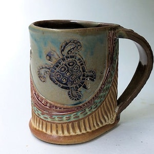 May include: A ceramic mug with a brown handle and a light brown, green, and blue glaze. The mug features a raised design of a sea turtle with a blue and black shell.