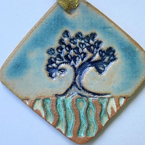 May include: A ceramic ornament with a tree design. The ornament is diamond-shaped with a blue and tan background. The tree is dark blue with detailed leaves and a textured trunk. The bottom has a brown and green wavy pattern. A gold ribbon is attached.