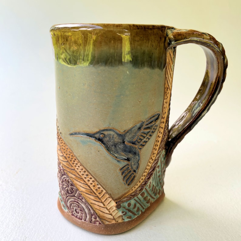 May include: A ceramic mug with a brown handle and a green, brown, and blue glaze. The mug features a raised design of a hummingbird in flight.