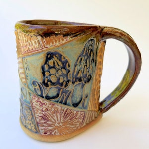 Può includere: Una tazza in ceramica fatta a mano con una glassa marrone, verde e blu. La tazza ha un design unico con un motivo a patchwork e un'immagine in rilievo di funghi.