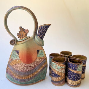 Può includere: Una teiera in ceramica e sei tazze abbinate con un design astratto colorato. La teiera ha un'unica maniglia curva e un coperchio decorativo. Le tazze sono piccole e hanno un design simile a quello della teiera.