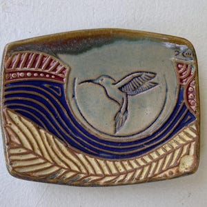 Puede incluir: Plato rectangular de cerámica con diseño de colibrí. El plato presenta un fondo azul claro con un colibrí en el centro. Los bordes tienen un borde marrón, con patrones de olas azules y elementos decorativos rojos y amarillos. El plato mide aproximadamente 15 cm de largo.