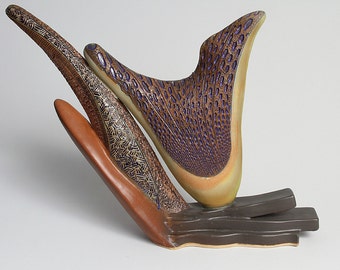 May include: A ceramic sculpture with three abstract forms. The largest form is a brown, yellow, and purple wing-like shape. The other two forms are long and thin, with a brown and gold color scheme.