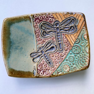 May include: A rectangular ceramic tray featuring a smooth, light blue glazed section on the left and a textured, multi-colored right side. The right side displays two embossed dragonfly figures surrounded by intricate circular and striped patterns.