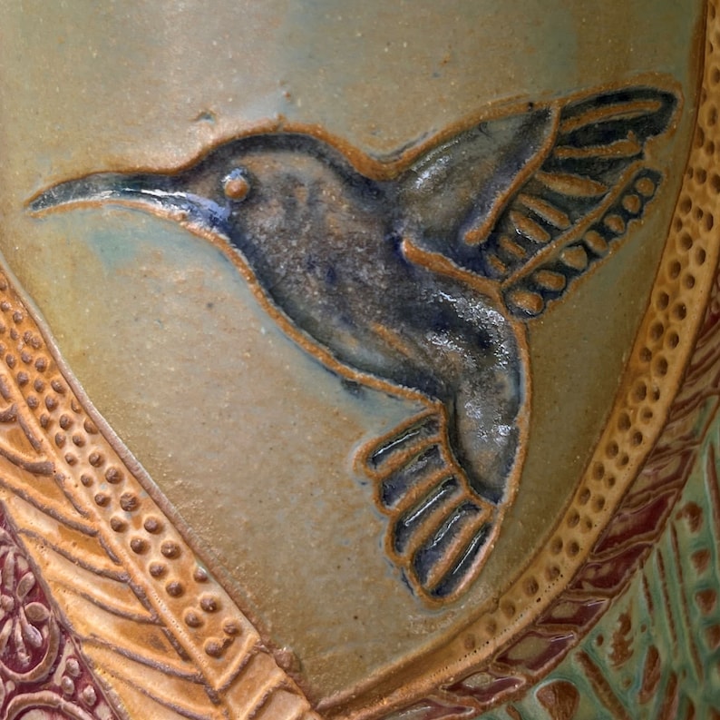 May include: A ceramic vase with a blue and brown hummingbird design. The vase has a textured surface with a brown and gold pattern.