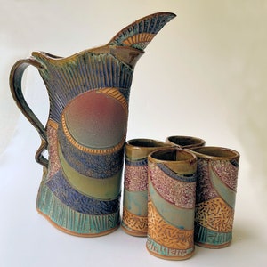 May include: A set of four brown, blue, and green ceramic shot glasses with a decorative design, arranged in a clover shape, and a matching brown, blue, and green ceramic pitcher with a decorative design.
