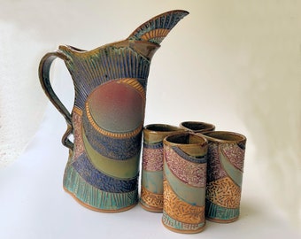 May include: A set of four brown, blue, and green ceramic shot glasses with a decorative design, arranged in a clover shape, and a matching brown, blue, and green ceramic pitcher with a decorative design.