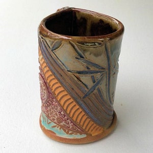 May include: A handmade ceramic mug with a colorful, patterned design. The mug features a brown glaze with orange, blue, and pink accents. The design includes geometric shapes and floral motifs.
