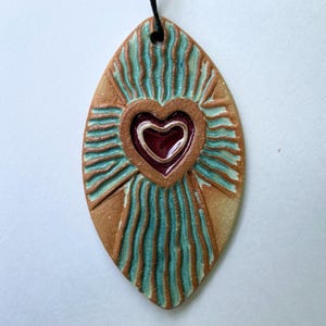 Könnte beinhalten: Handgefertigter Keramikanhänger mit Herzdesign. Der Anhänger hat eine längliche ovale Form mit einem zentralen Herzen in tiefem Rot, umgeben von einer braunen Herzform. Der Hintergrund hat türkisfarbene Streifen und einen beigen Rand. Eine schwarze Kordel ist befestigt.