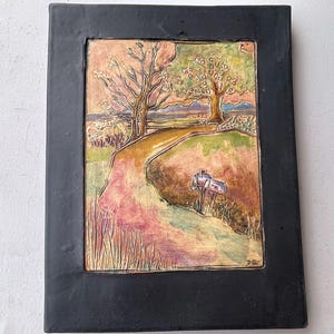 May include: A framed ceramic artwork depicting a rural landscape with a winding road, trees, and a mailbox. The artwork features a color palette of pinks, greens, and browns, set within a black frame. The artwork is approximately 12 inches tall and 9 inches wide.