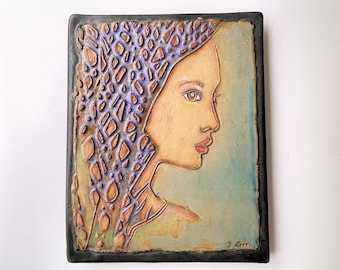 May include: A ceramic tile with a raised relief image of a woman's face. The woman is wearing a patterned head covering with purple and brown accents. The background is a light blue-green color.