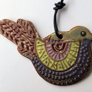 Könnte beinhalten: Ein Keramik-Vogel-Anhänger mit einem farbenfrohen, geometrischen Design. Der Vogel hat einen grünen Kopf, einen violetten Körper und gelbe Flügel. Er ist mit braunen, roten und weißen Akzenten verziert. Der Vogel hängt an einer schwarzen Schnur.