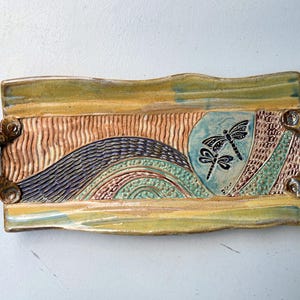 May include: A ceramic serving tray with two dragonfly designs and a wavy, textured pattern in shades of brown, green, blue, and yellow. The tray has two handles on the sides.