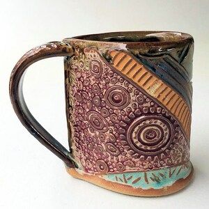 May include: A handmade ceramic mug with a brown, orange, blue, and white geometric design. The mug has a textured surface with a pattern of circles and dots.