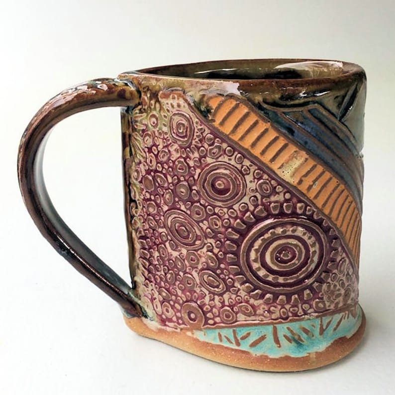 May include: A handmade ceramic mug with a brown, orange, blue, and white geometric design. The mug has a textured surface with a pattern of circles and dots.
