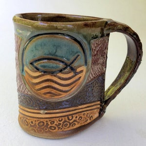 May include: A handmade ceramic mug with a brown, green, and blue glaze. The mug features a fish and waves design in the center. The mug has a textured surface and a handle.