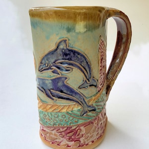May include: A ceramic mug with a handle, featuring a hand-painted design of two dolphins swimming in a blue and green ocean scene. The mug has a brown, green, and red glaze.