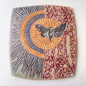 May include: Square ceramic wall art featuring a moth design. The artwork is divided into two sections: one with a sunburst pattern and the other with a textured, reddish-brown surface. The moth is blue and orange.