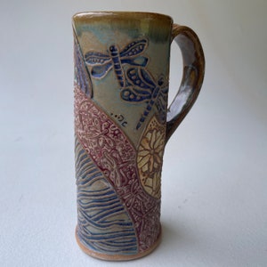 Peut inclure: Une tasse en céramique avec un glaçage bleu, vert et brun. La tasse a une anse et est décorée d'un motif de libellule.