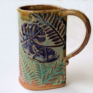May include: A handmade ceramic mug with a brown, green, and purple leafy design. The mug has a wide, rounded base and a handle.