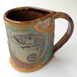 May include: A ceramic mug with a brown, green, and blue glaze. The mug features a detailed design of a chameleon with a curled tail. The handle is brown and slightly curved.
