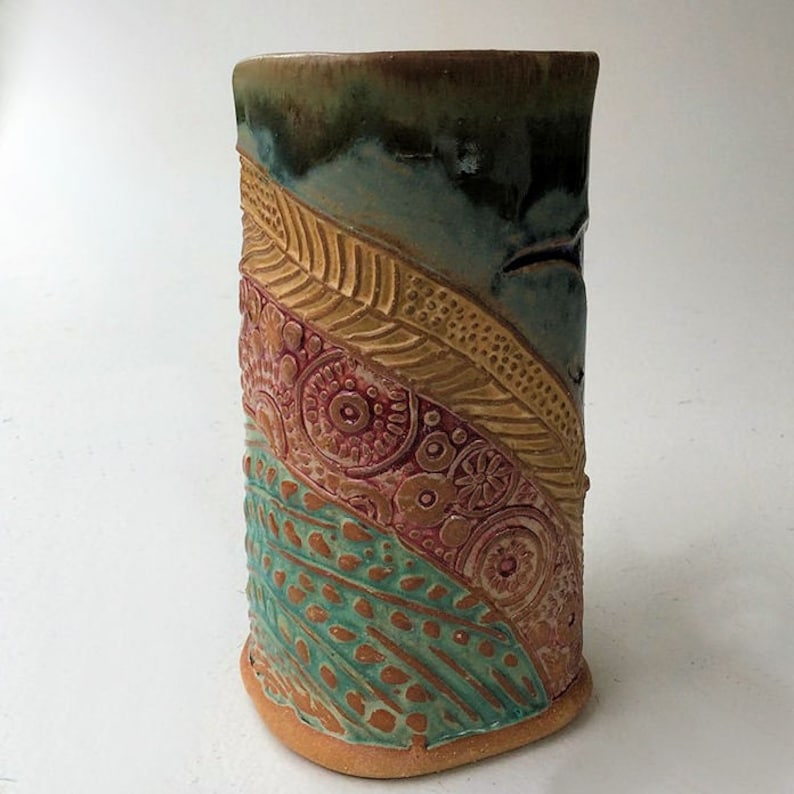 May include: A ceramic vase with a textured, multi-colored design. The vase is decorated with a pattern of leaves, circles, and dots in shades of brown, yellow, green, and red.