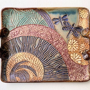 May include: A ceramic serving tray with two sculpted handles. The tray features a colorful, abstract design with swirls, lines, and floral patterns in shades of blue, green, brown, and pink. Two dragonfly designs are incorporated into the pattern.