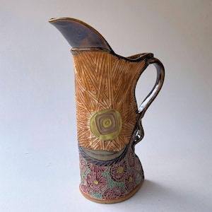 Op de afbeelding: Handgemaakte keramische kan met een getextureerd, bruin oppervlak. Het ontwerp omvat een zonnepatroon en bloemendetails aan de basis. De handgreep en tuit zijn donkerbruin. De kan is ongeveer 25 cm hoog.