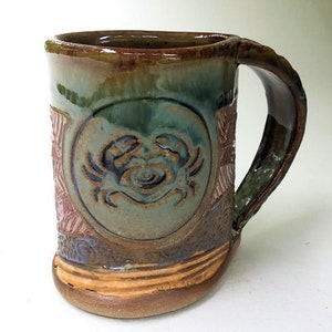 May include: A handmade ceramic mug with a brown, green, and purple glaze. The mug features a raised design of a crab in a circle on the front.
