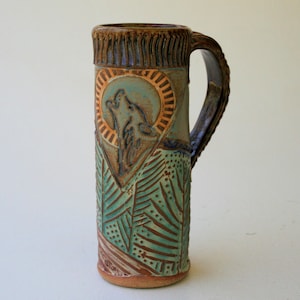 May include: A ceramic mug with a brown, green, and blue design. The mug features a wolf howling at the moon, surrounded by a sunburst design. The mug has a handle and is approximately 15 cm tall.