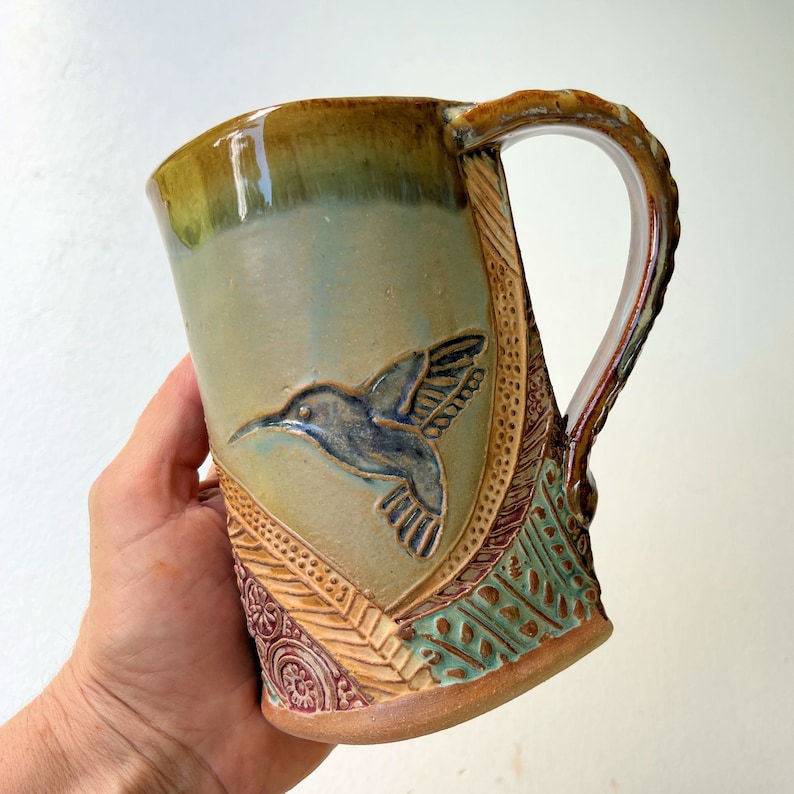 May include: A handmade ceramic mug with a brown, green, and blue glaze. The mug features a detailed hummingbird design and intricate patterns around the rim.