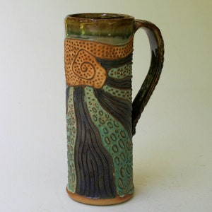 May include: A tall, ceramic mug with a brown, green, and orange design. The mug has a large handle and a textured surface.