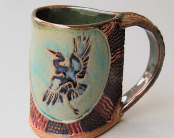 May include: A ceramic mug with a blue and black bird design on a teal background. The mug has a brown handle and is decorated with red, brown, and black patterns.