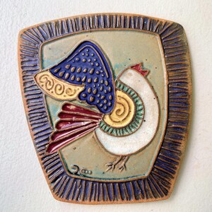 May include: A ceramic tile featuring a stylized bird design. The bird has a white body, a blue wing with a dotted pattern, and a red and yellow tail. The tile is framed with a dark blue border and has a light green background.