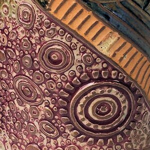 May include: Close-up of a ceramic vase with a textured surface. The vase has a brown, orange, and purple design with a repeating pattern of circles and dots.