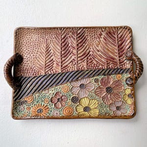 May include: Rectangular ceramic tray with a textured surface and two handles. The tray features a floral design with brown, yellow, and orange flowers. The upper section has a dotted pattern and leaf-like designs in shades of brown and tan.