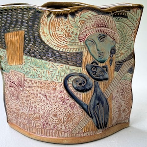 May include: A ceramic vase with a unique design. The vase features a woman's face, a black cat, and various patterns in shades of blue, green, brown, and tan. The vase has a textured surface and a curved shape, adding to its artistic appeal.