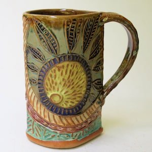May include: A handmade ceramic mug with a green, brown, and yellow glaze. The mug has a unique design with a circular pattern and leaf-like shapes.