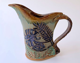 May include: A brown and green ceramic pitcher with a unique, curved handle. The pitcher is decorated with a fern and leaf design in shades of blue, green, and brown.