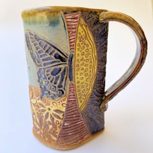 May include: A handmade ceramic mug with a butterfly design. The mug is decorated with a blue butterfly, yellow and red geometric patterns, and brown floral designs.